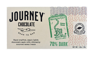 A medium size packaged Journey Chocolate bar in an off-white cardboard box with wavy US postal service lines across the top and right sides of the box. A white postage stamp style seal with a cacao pod in the middle seals the box, with the words "Experts in small-batch, craft chocolate" in a circle around the image and the words "Seal of Quality" at the bottom. The journey chocolate logo is on the left side of the  bar and an Madagascar 70% dark stamp centered in the middle, net weight 1.0 oz or 28 grams.